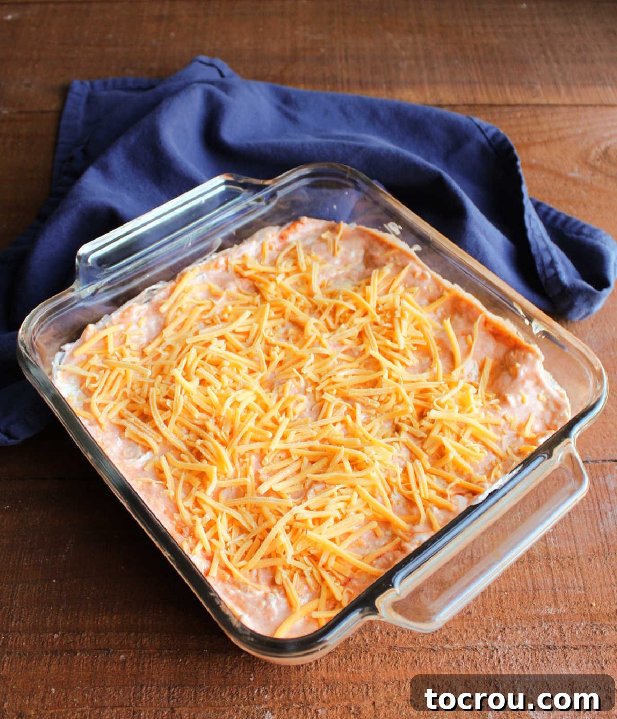 Ultimate Creamy Buffalo Chicken Dip 5 The buffalo chicken dip mixture, creamy and orange, is transferred into a clear glass square baking dish, perfectly smoothed and ready to be topped with the remaining cheese before entering the oven.