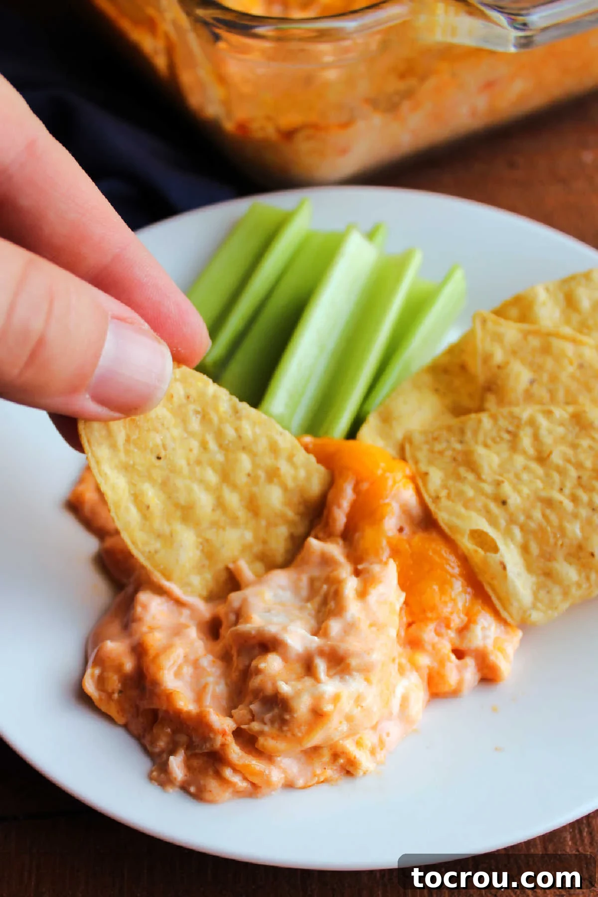 Ultimate Creamy Buffalo Chicken Dip 8 A hand scooping a crispy tortilla chip into a freshly served portion of warm buffalo chicken dip, illustrating the perfect creamy and chunky texture for dipping.