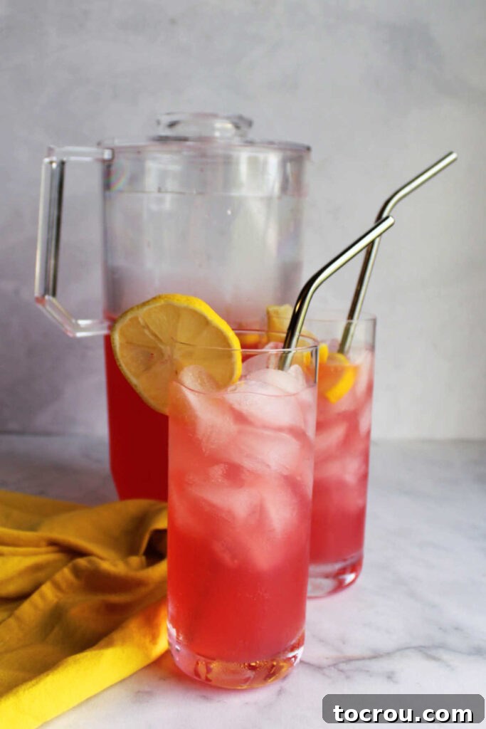 Large glass pitcher of freshly made pink lemonade with ice and slices of lemon.