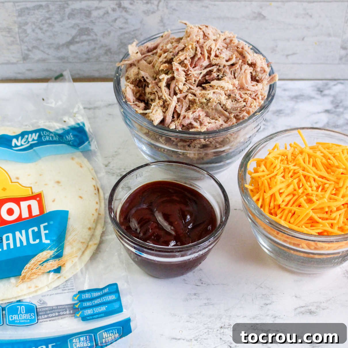 Pulled Pork Taquito Ingredients Ingredients laid out: shredded pulled pork, a bowl of BBQ sauce, a pile of shredded cheese, and a stack of tortillas, ready for taquito assembly.