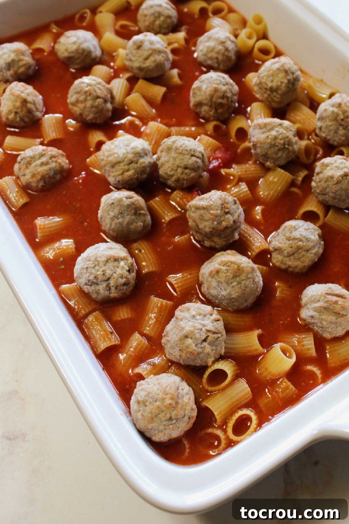 Uncooked pasta and tomato sauce in dish with frozen meatballs ready for the oven.