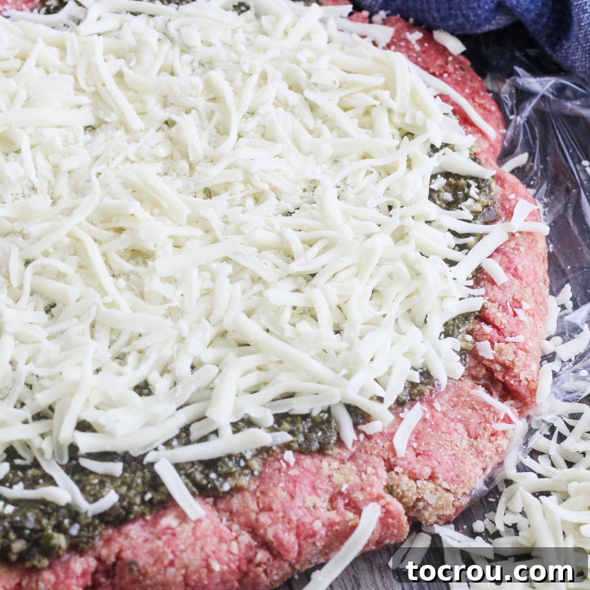 Layer of ground beef covered with pesto and cheese ready to be rolled into meatloaf.