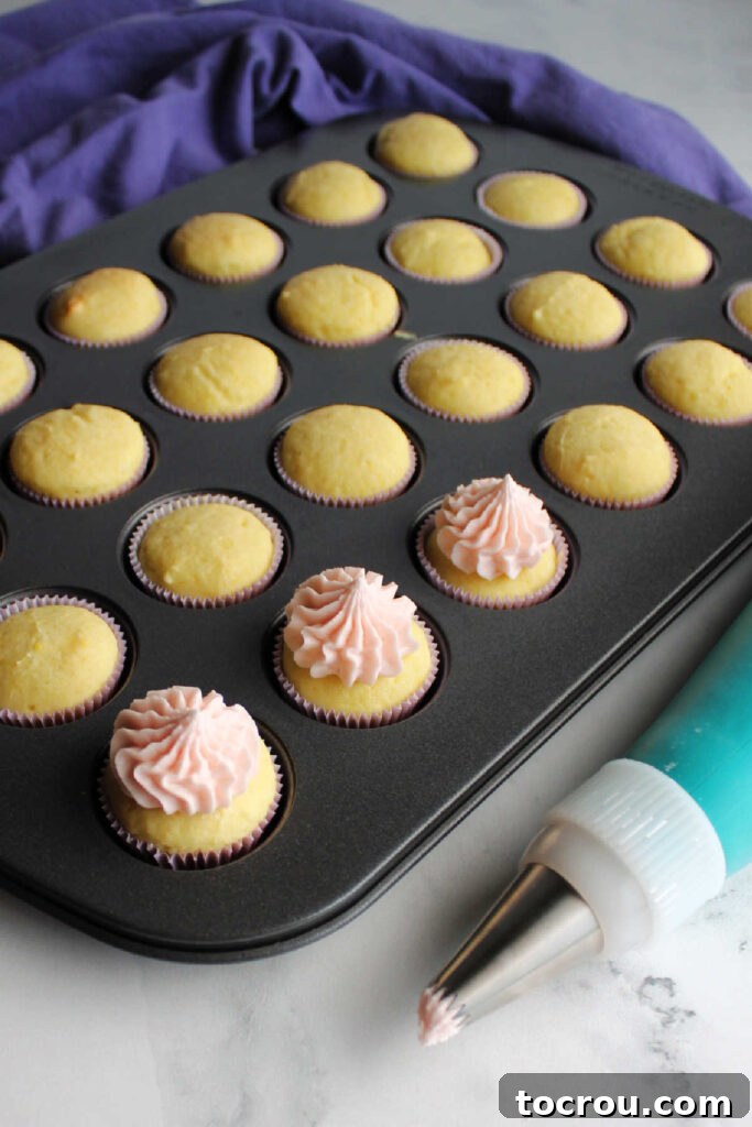 Piping bag next to a pan of mini cupcakes, some adorned with pink lemonade frosting.