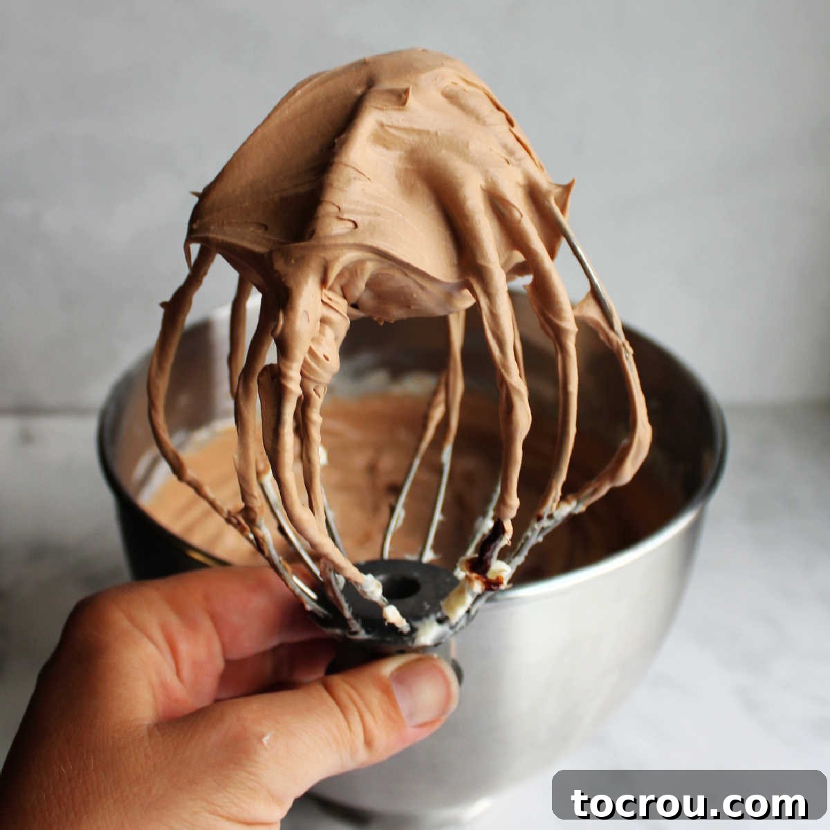 A mixer whisk filled with fluffy, light brown chocolate cheesecake mixture, ready to be piped.