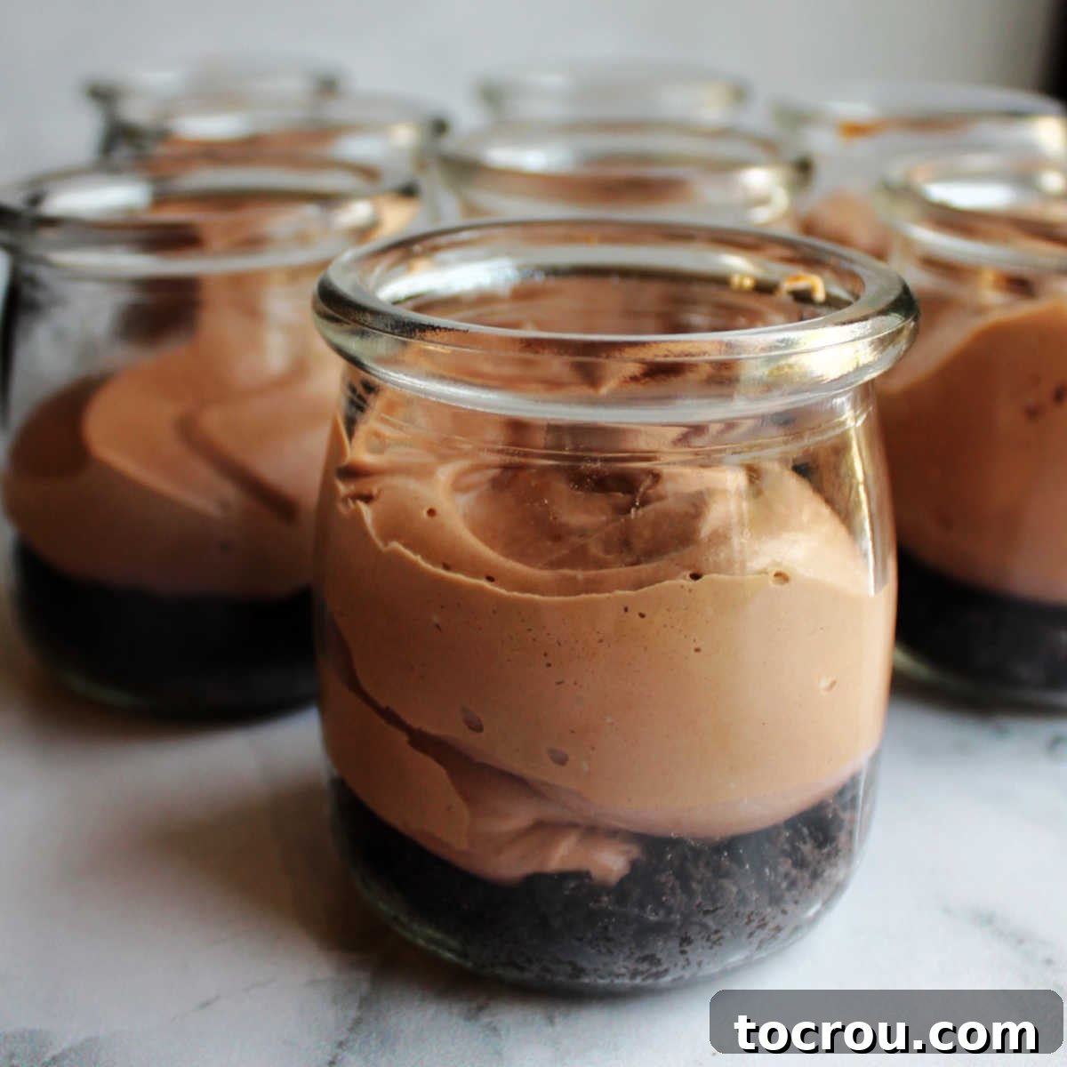Rows of small jars with chocolate cookie crumb crusts at the bottom, topped with freshly piped creamy chocolate cheesecake mixture.
