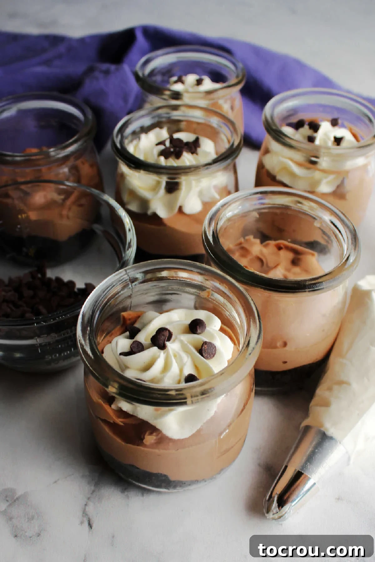A piping bag elegantly adding a swirling layer of cream cheese whipped cream onto the tops of chocolate cheesecake jars.