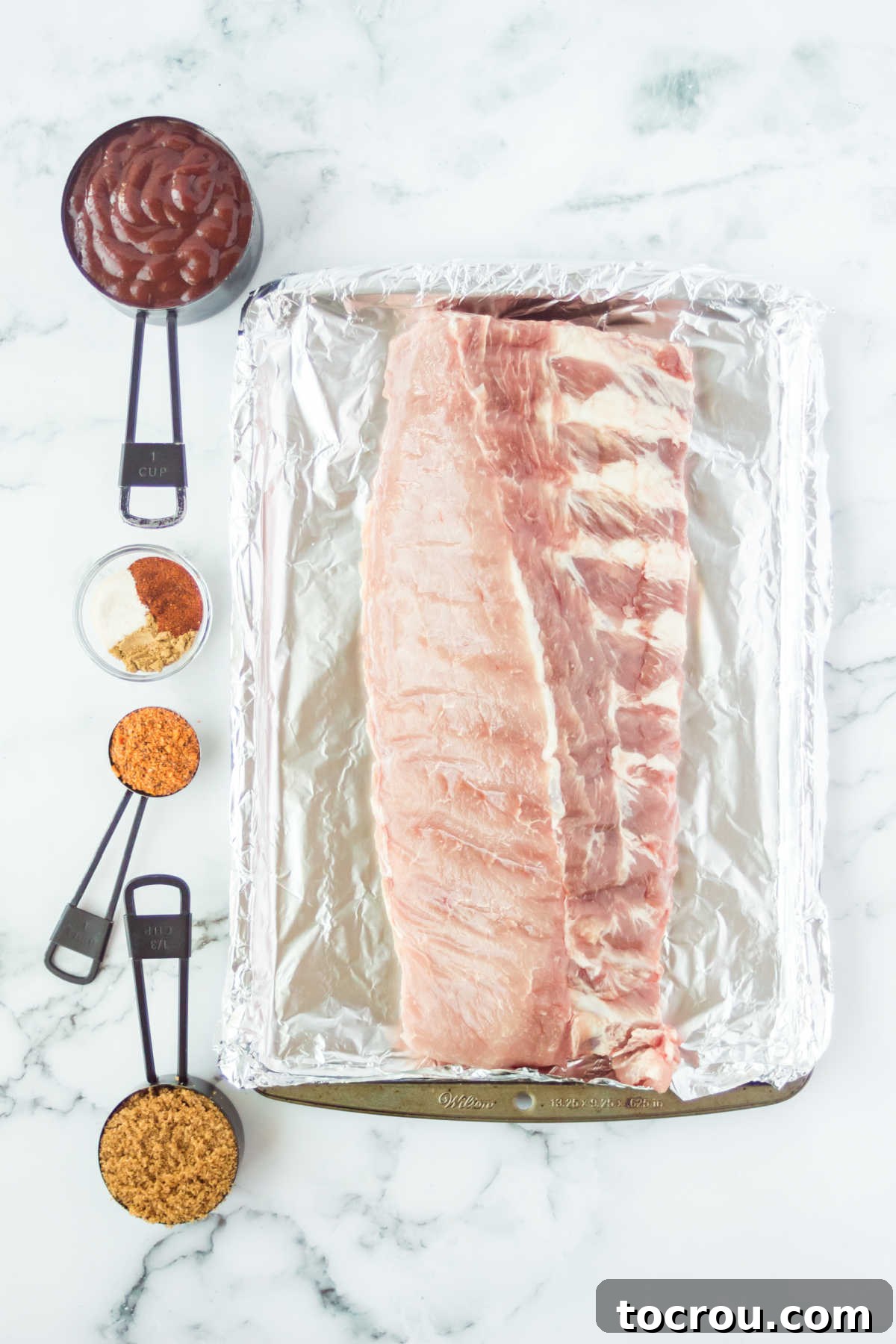 Mouthwatering Oven Baked Ribs 3 Rack of ribs on foil lined pan with seasonings and sauce nearby.