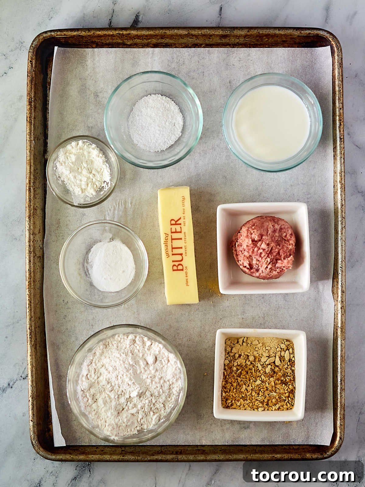 Fluffy Biscuits and Rich Sausage Gravy 4 Various ingredients laid out on a surface, including flour, salt, milk, cubes of butter, ground sausage, and assorted seasonings, all prepared for making homemade biscuits and gravy.