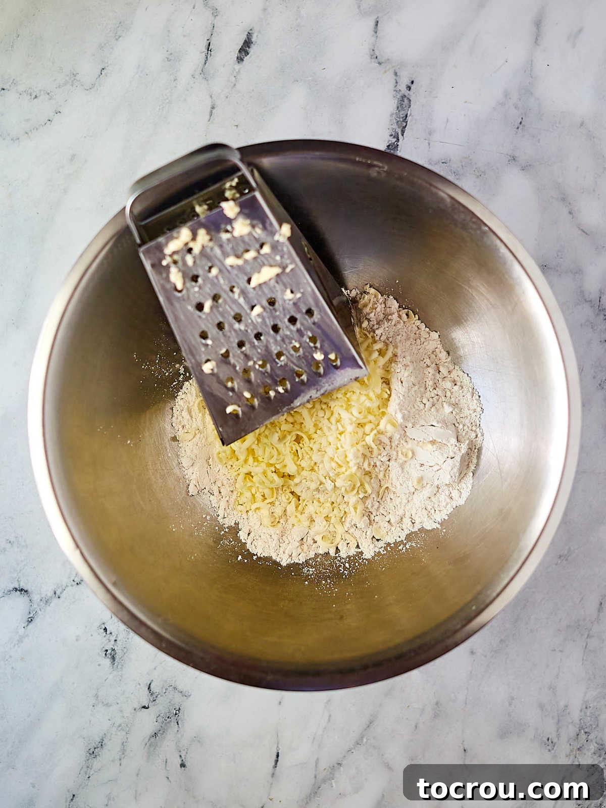 Fluffy Biscuits and Rich Sausage Gravy 5 A mixing bowl containing a flour mixture, with a grater resting on the edge and shredded frozen butter sprinkled over the flour.
