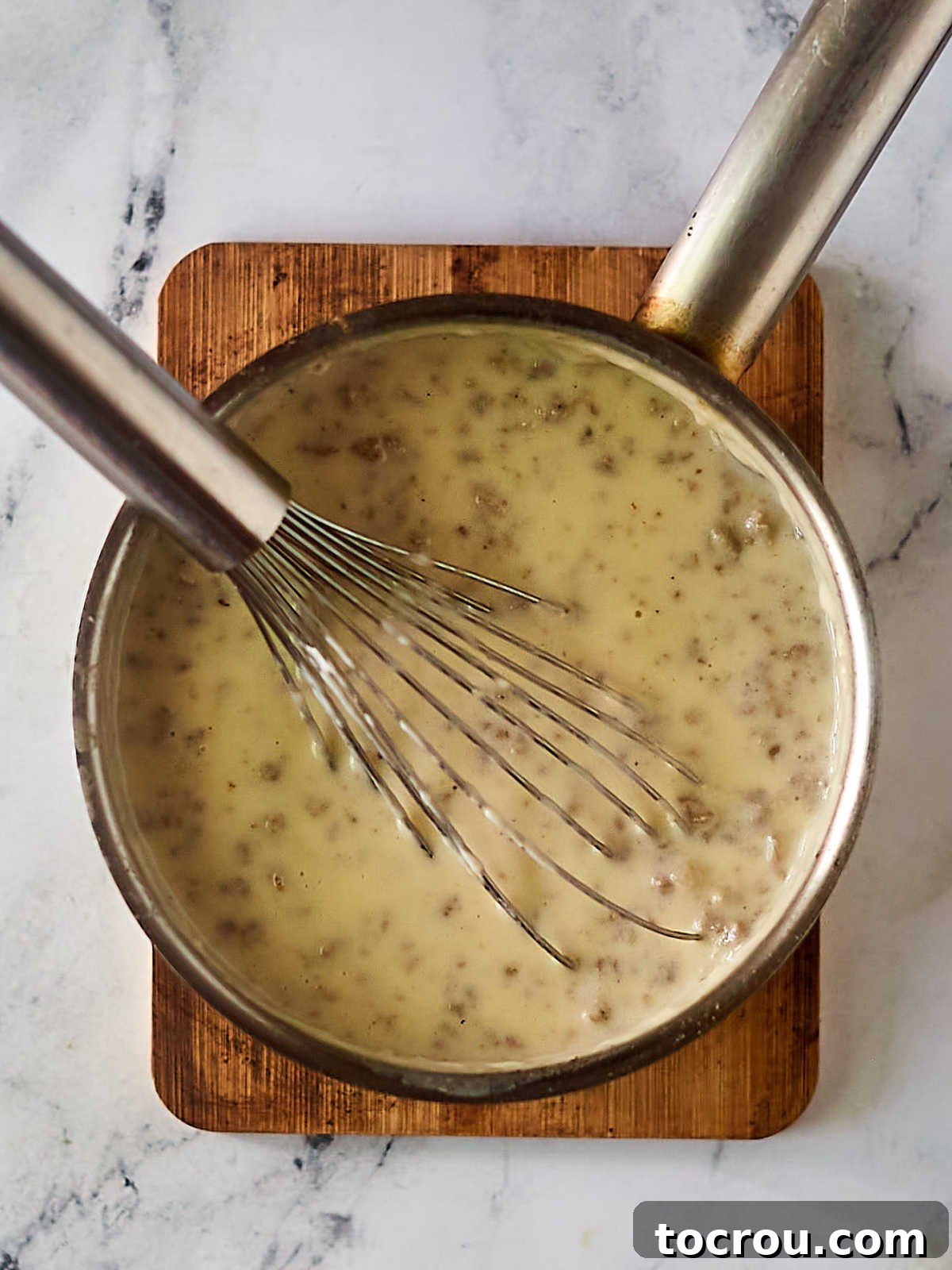 Fluffy Biscuits and Rich Sausage Gravy 10 Sausage gravy in a skillet, having just had milk added and cooked until visibly thickened, with a whisk resting in it, ready to be served over biscuits.