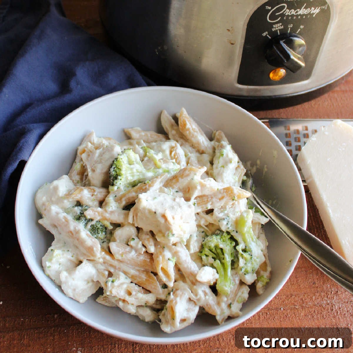 A delectable bowl of penne pasta with succulent chicken, fresh broccoli florets, and a generous pour of creamy alfredo sauce, next to a slow cooker.