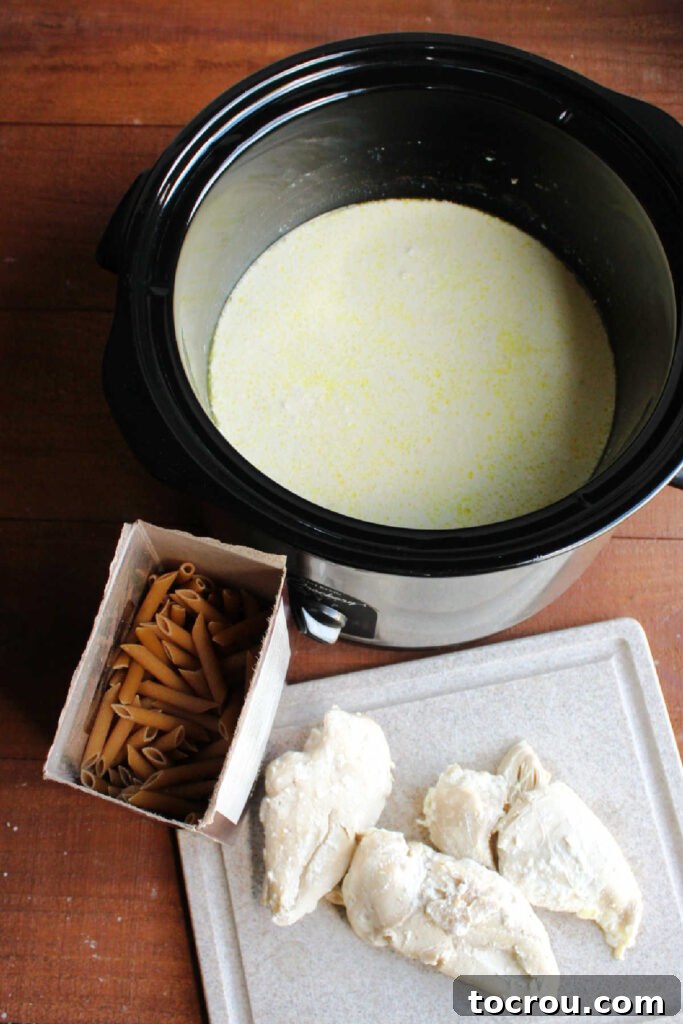 Tender, cooked chicken breasts on a cutting board, ready for shredding, next to a slow cooker filled with the creamy alfredo sauce base.