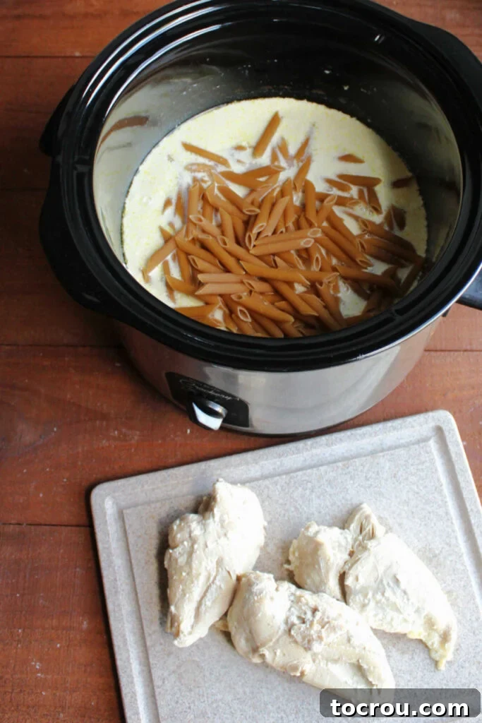 Uncooked penne pasta being added to the slow cooker, which holds the creamy alfredo sauce and cooked chicken.