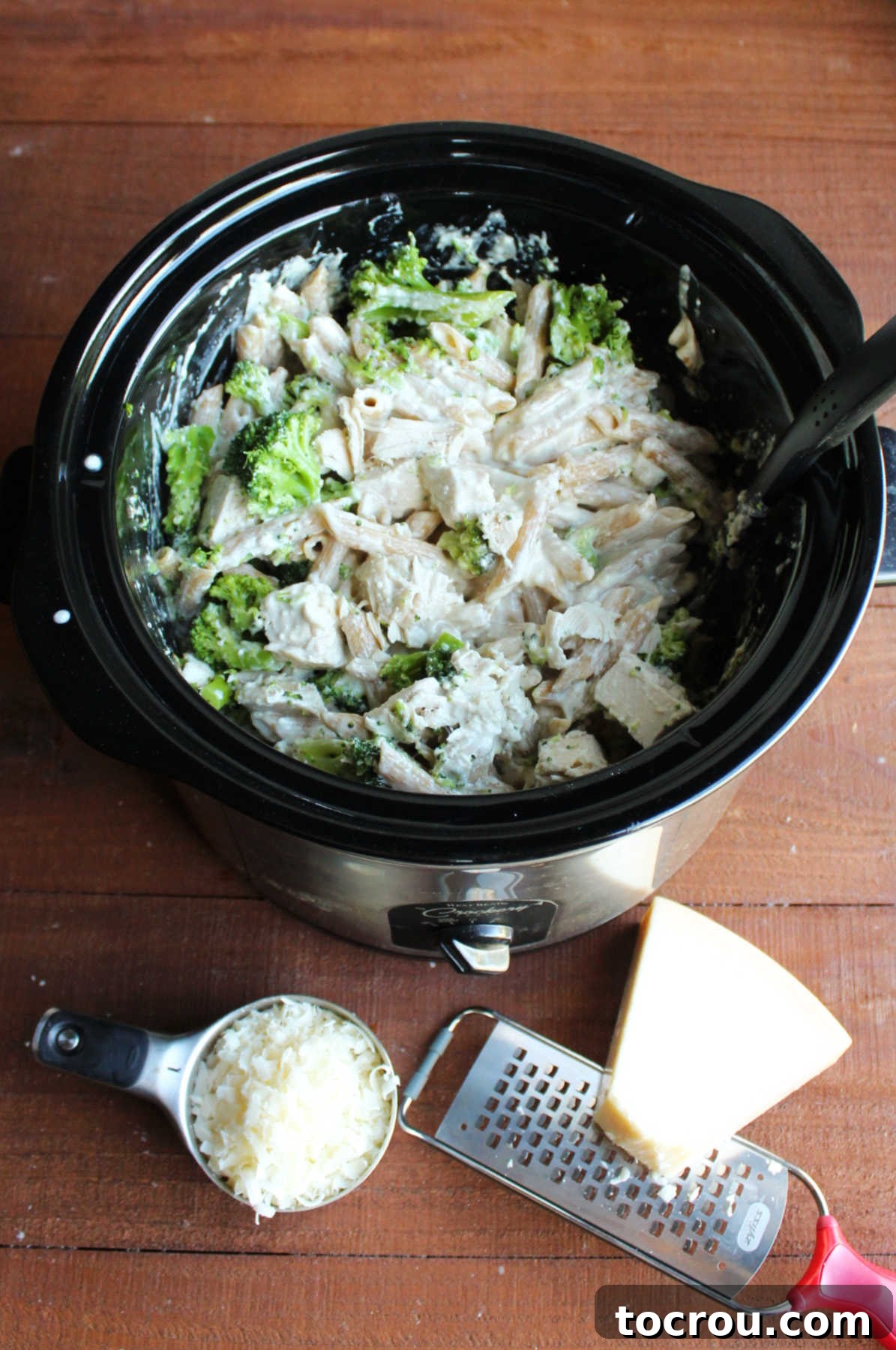 A hand grating fresh Parmesan cheese over the finished slow cooker chicken alfredo, with pasta, chicken, and broccoli visible.