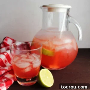 Pitcher of pink cherry limeade next to glass of limeade and a lime half.