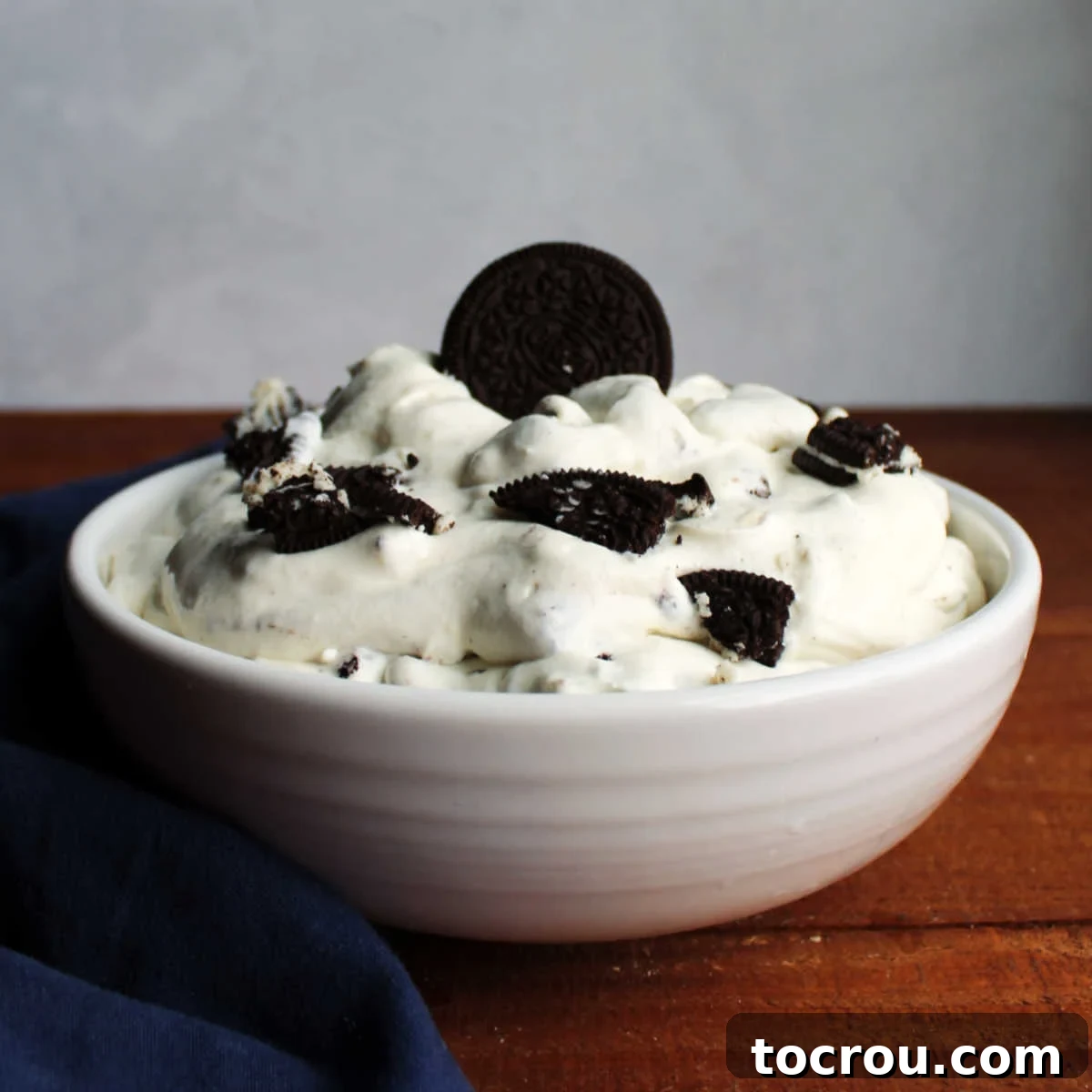 Delicious 4-Ingredient Oreo Fluff Dessert A large glass bowl overflowing with creamy Oreo fluff salad, generously topped with crumbled cookie pieces and one whole chocolate sandwich cookie as a garnish. The texture is light and airy.
