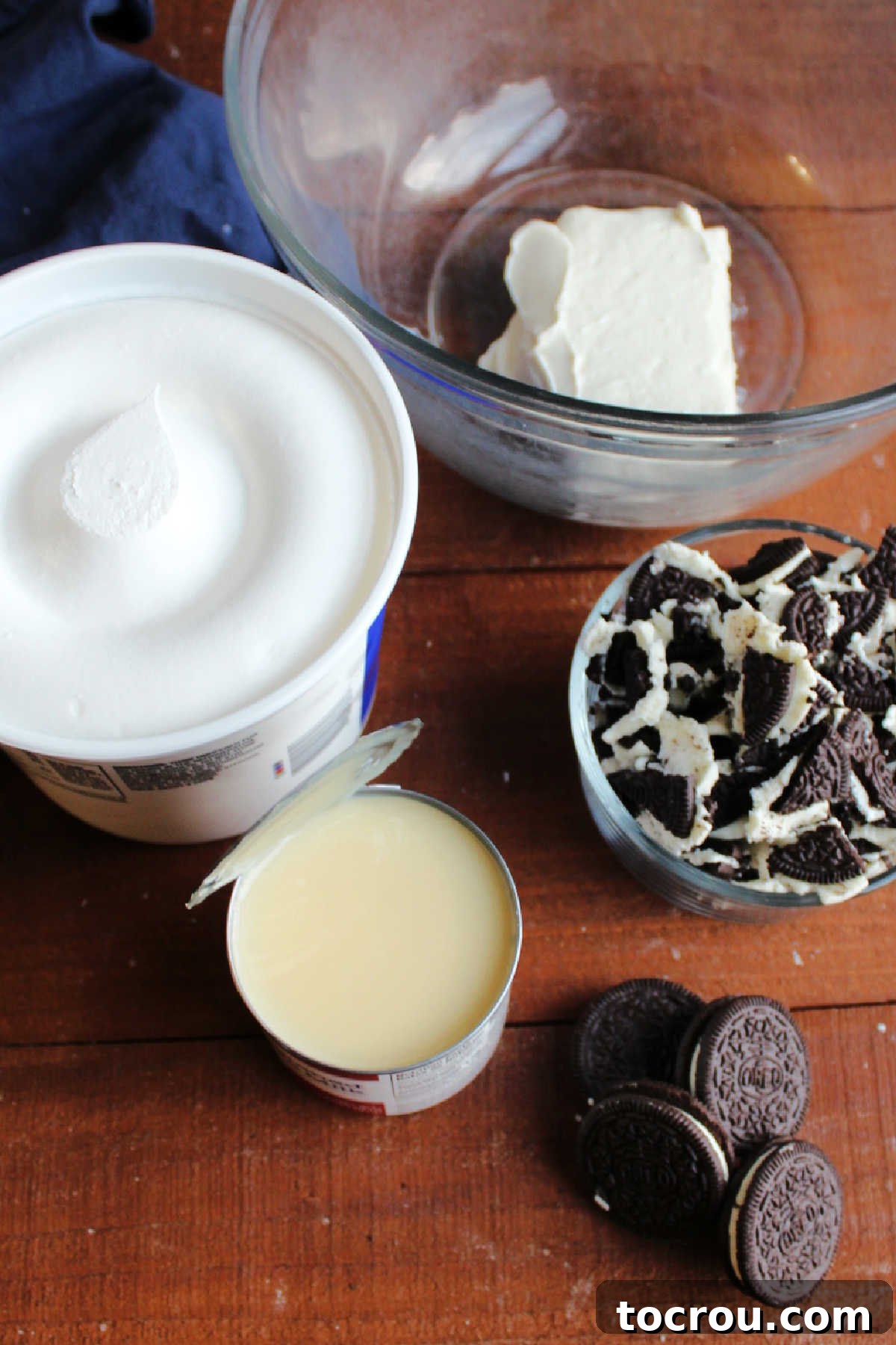 Ingredients for Oreo Fluff Four primary ingredients laid out on a white surface: a block of cream cheese, a can of sweetened condensed milk, a tub of Cool Whip, and a package of Oreo cookies. All essentials for making the fluff.
