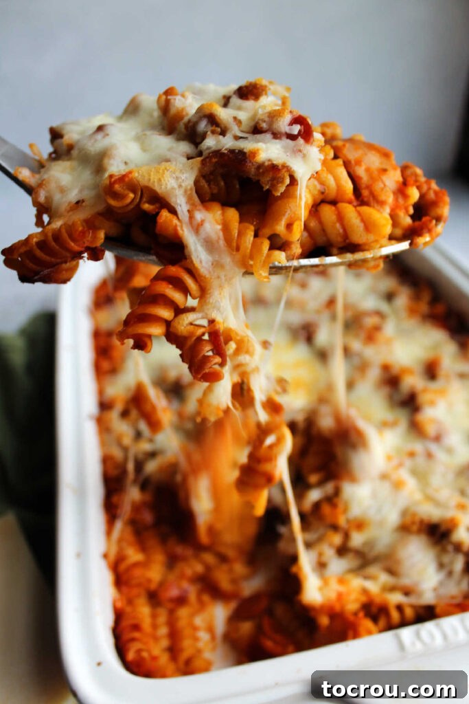 Serving spoon lifting out helping of pizza pasta bake with strings of melted mozzarella cheese pulling from dish.