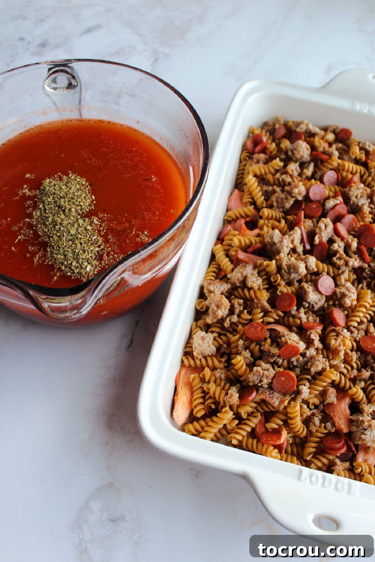 Pan of pasta topped with sausage and pepperoni next to bowl of sauce with oregano and garlic, ready for the oven.