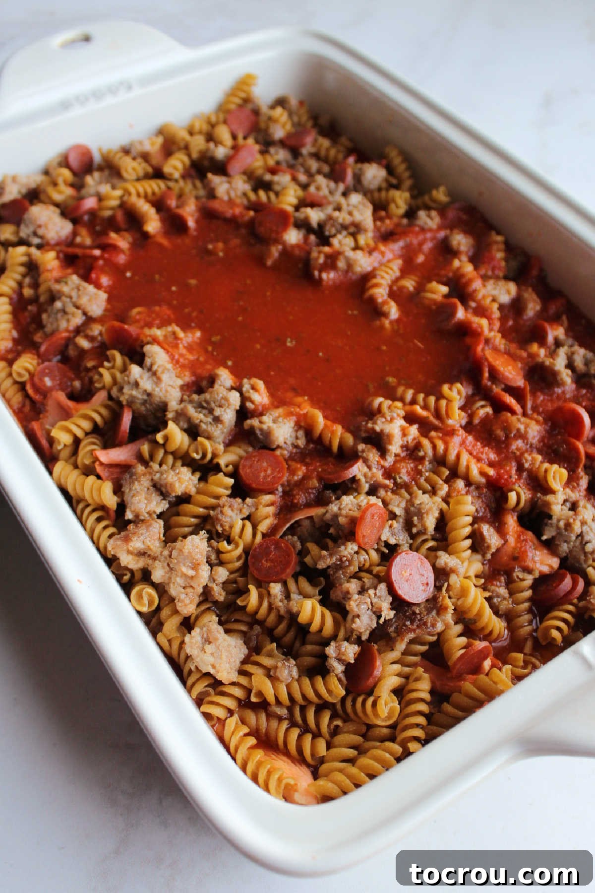 Raw pasta in pan with sausage, pepperoni and sauce mixture ready to go in the oven, illustrating easy prep.