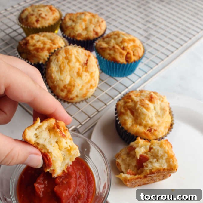 Savory Pepperoni Pizza Muffins dipped in marinara sauce Hand dipping a savory pepperoni pizza muffin into a small bowl of marinara sauce, with more freshly baked muffins visible in the background.