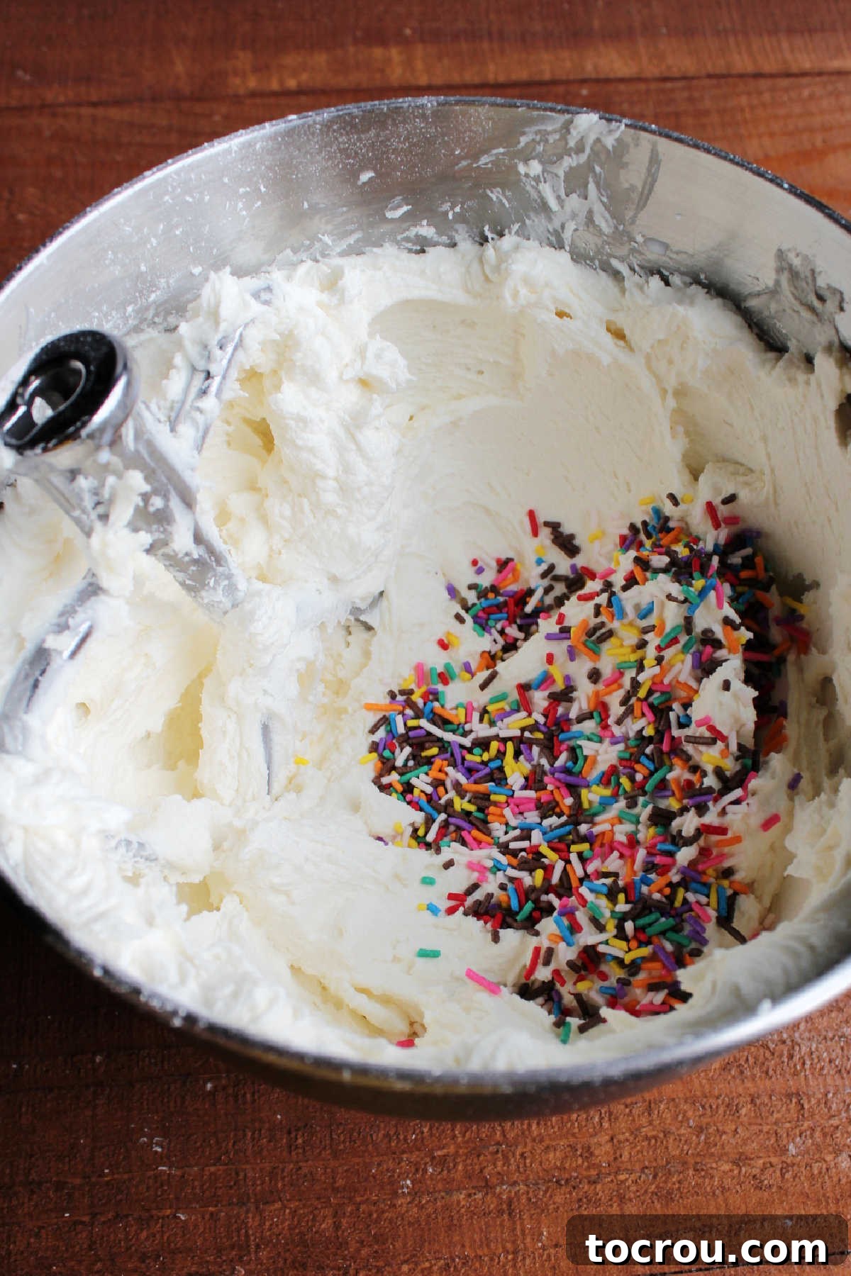 Bowl of buttercream with pile of sprinkles inside, ready to be mixed together.