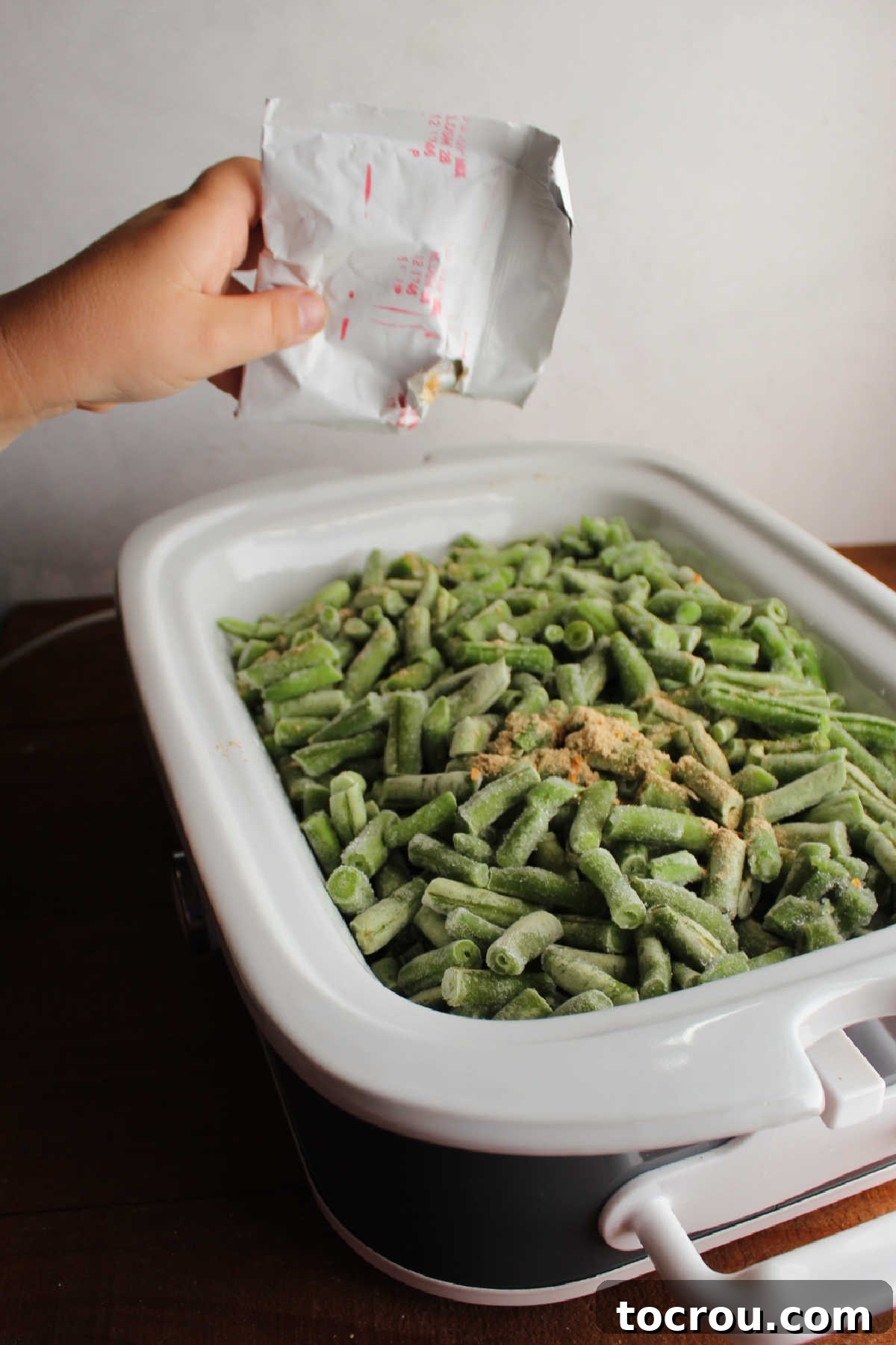 Child's hand sprinkling soup mix over slow cooker filled with frozen green beans.