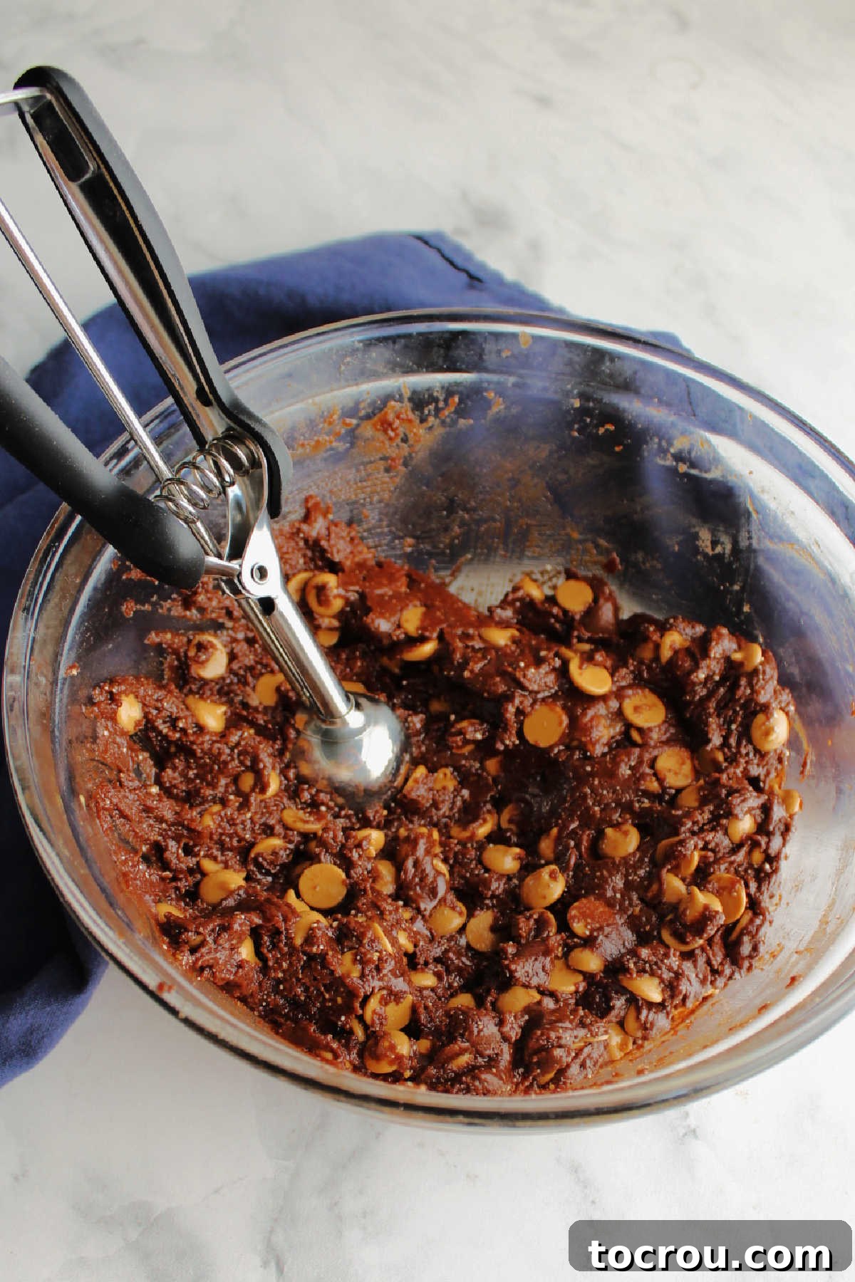 A bowl of rich chocolate cookie dough, generously mixed with peanut butter chips, alongside a cookie scoop, ready for portioning.
