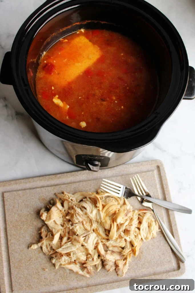 Shredded Chicken for Creamy Chili shredded chicken on cutting board in front of slow cooker with soupy broth.