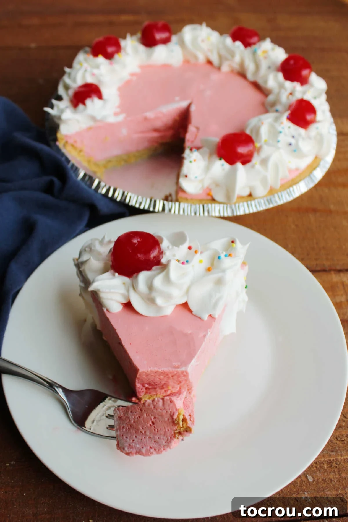 Bite of cherry jello on fork next to slice of pie in front of remaining pie, emphasizing the creamy texture.