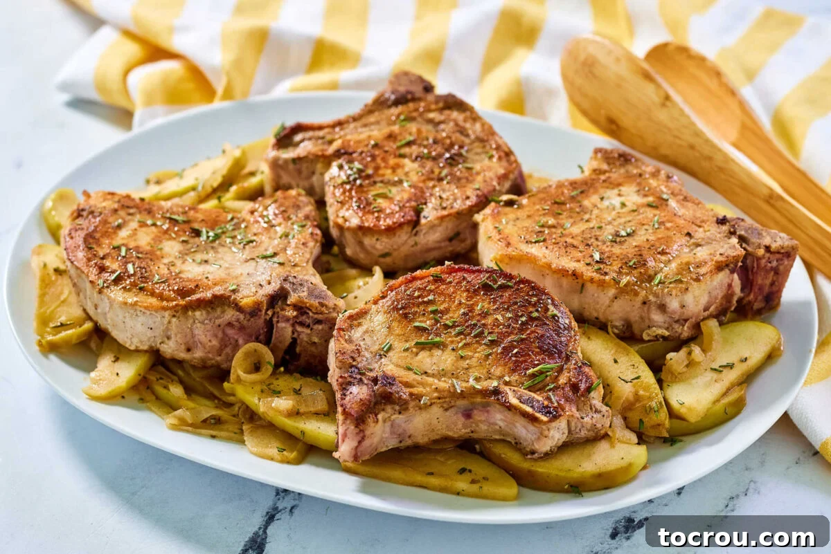 Platter of golden thick-cut pork chops nestled on a bed of cooked apples and onions, garnished with fresh rosemary, ready to serve.