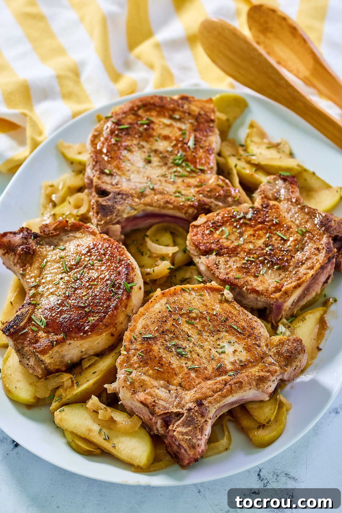 Platter of thick-cut bone-in pork chops presented on a mixture of cooked apples and onions, adorned with fresh rosemary garnish.