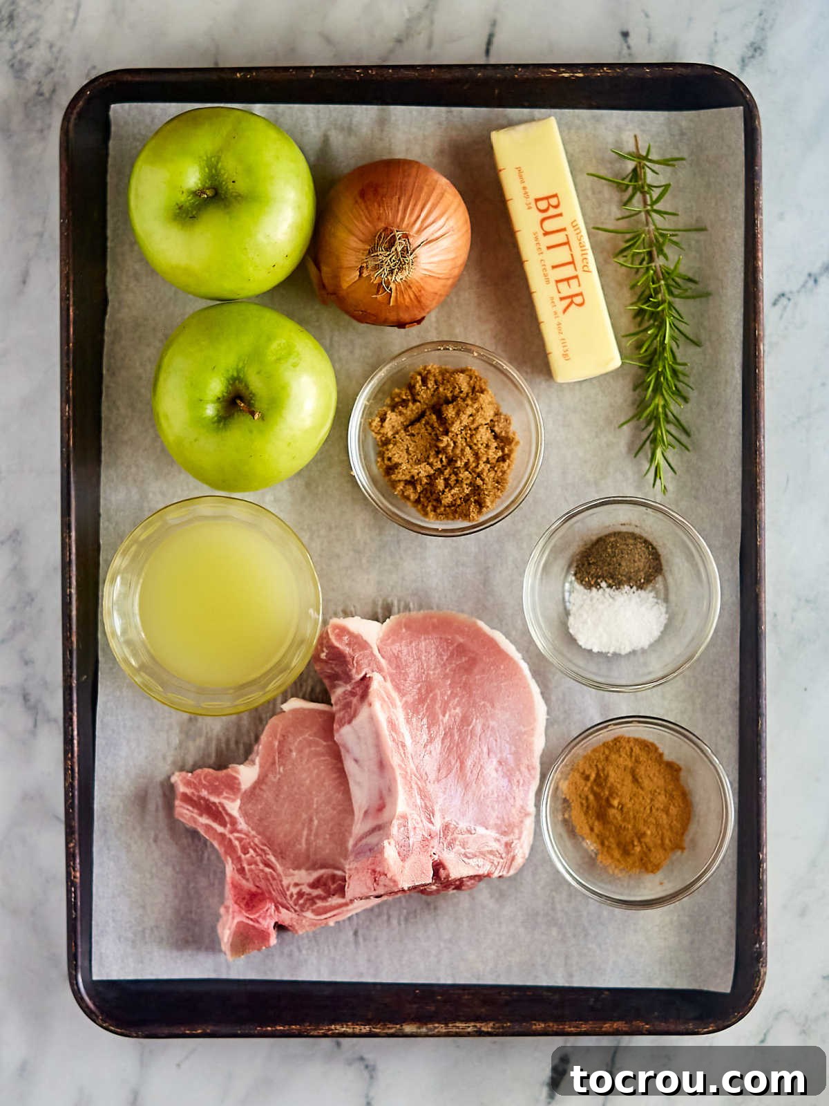 Ingredients for Skillet Pork Chops with Apples and Onions, including thick-cut bone-in pork chops, fresh apples, sliced onion, butter, fresh rosemary, and various seasonings, laid out before cooking.