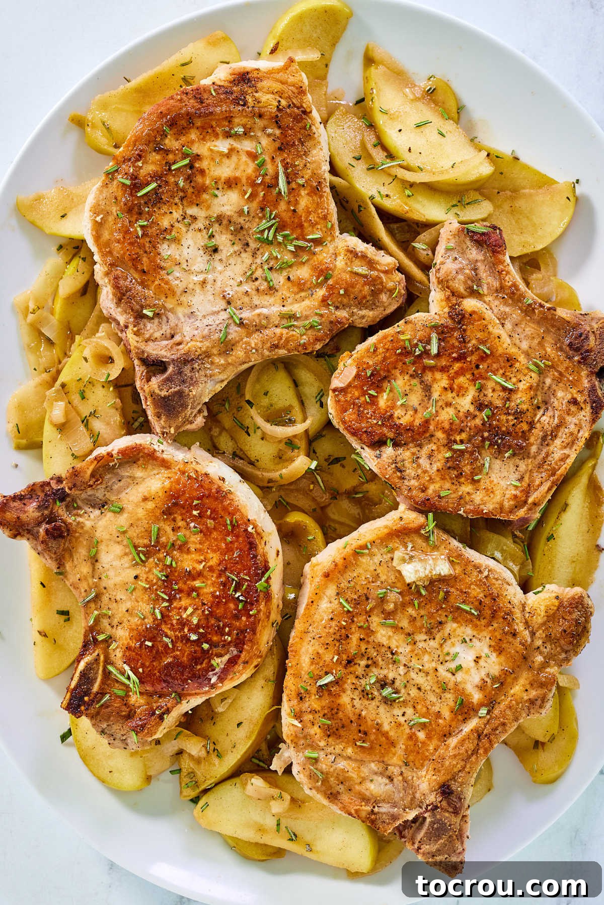 Close-up view of a bone-in pork chop topped with cooked apples and onions, garnished with fresh rosemary, ready to eat.