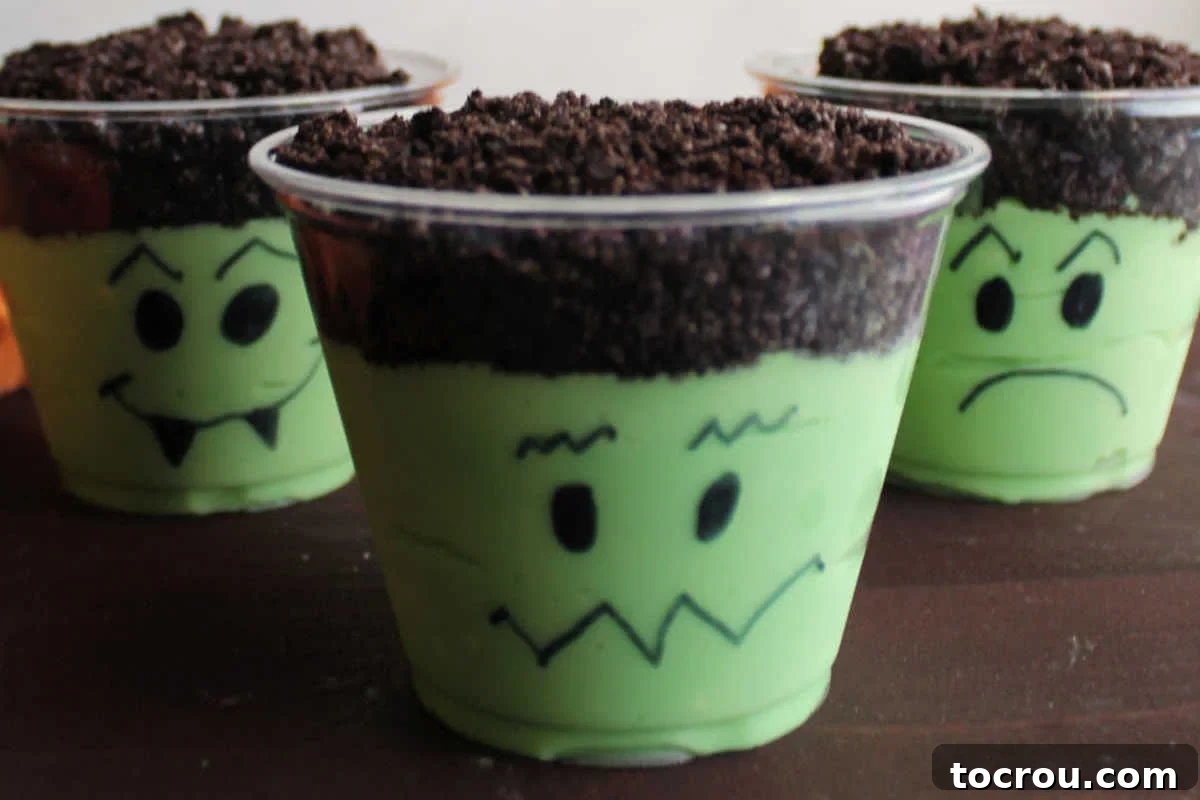 Three Frankenstein pudding cups with vibrant green pudding, topped with crushed Oreo 'hair,' and hand-drawn monster faces on the clear plastic cups.