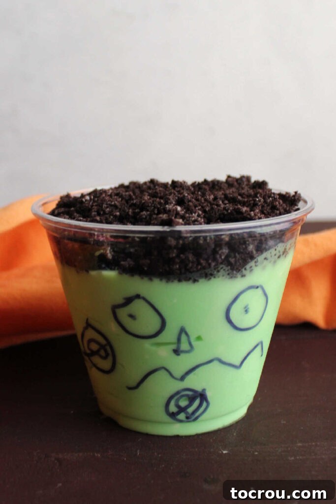 A close-up of a Frankenstein pudding cup with bright green pudding, a layer of crushed Oreos, and a childlike face drawn on the cup.