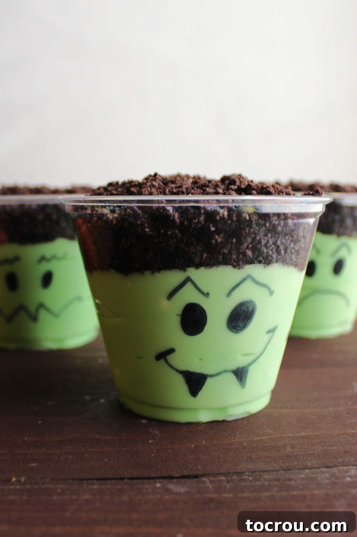A lineup of finished Frankenstein pudding cups, each with green pudding, Oreo hair, and unique monster faces drawn on the cups, ready for a Halloween party.