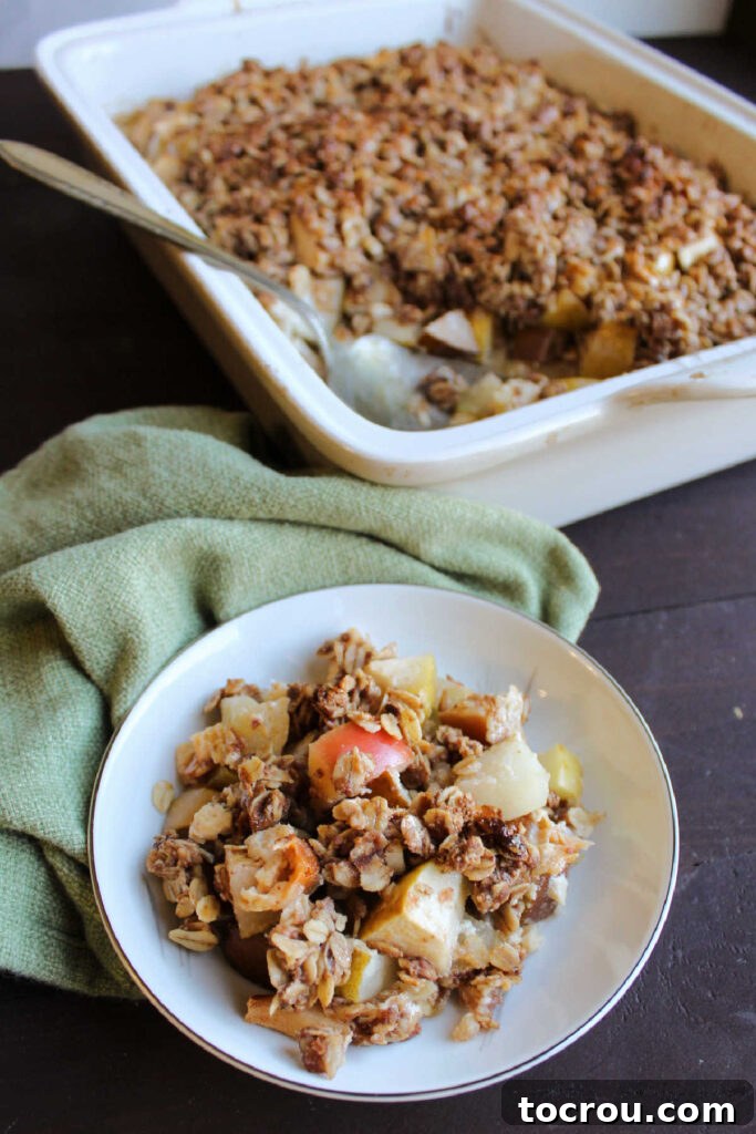 Serving of apple crisp on small plate in front of pan of remaining crisp.