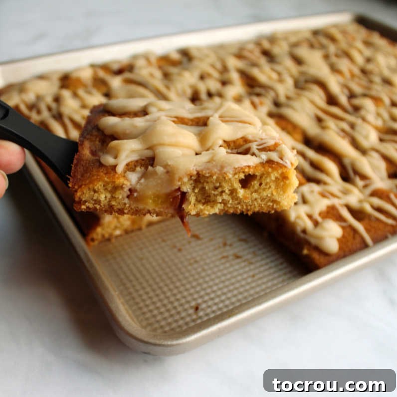 Summer Peach Sheet Cake 2 Spatula lifting a generous slice of peach sheet cake from the pan, revealing succulent fruit chunks baked into the moist crumb and a glistening drizzle of browned butter icing on top.