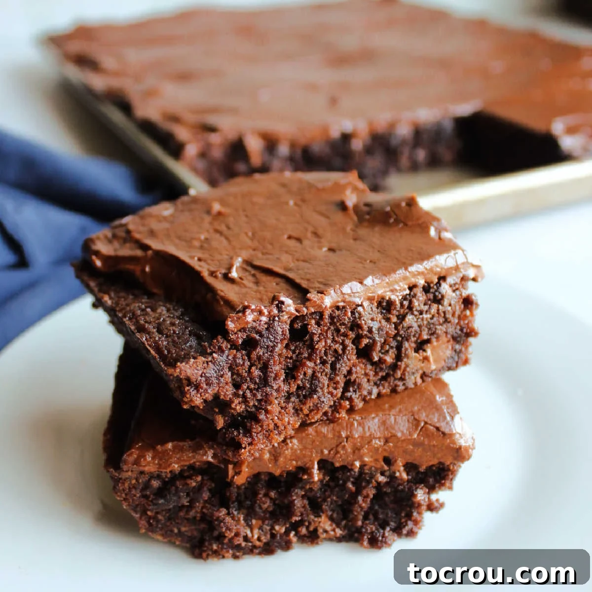 Classic Lunch Lady Brownies Two frosted lunch lady brownies stacked on top of each other with sheet pan of brownies in the background.