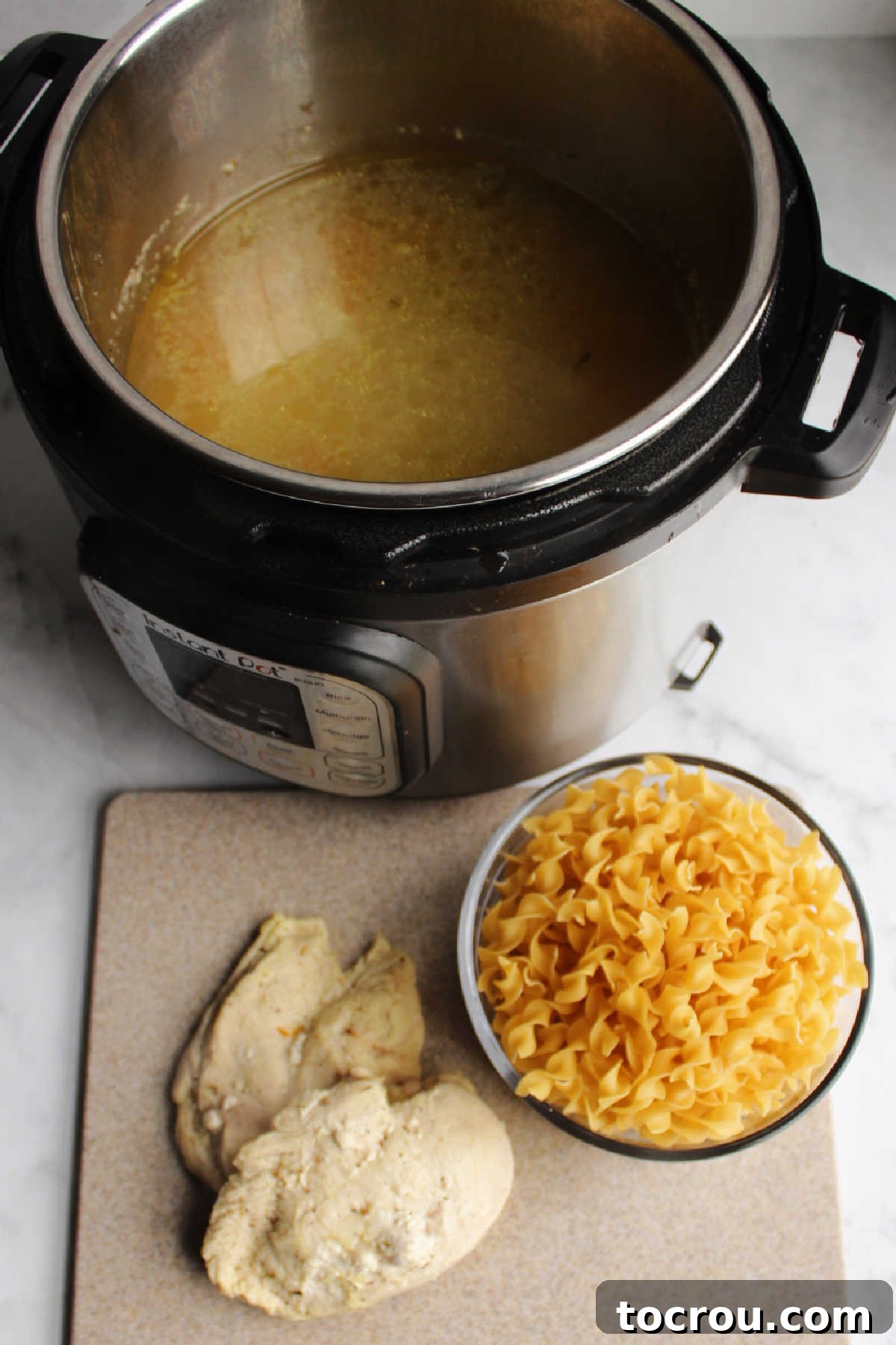 Flexible Chicken Noodle Soup: Instant Pot and Slow Cooker Ready 4 Cooked, shredded chicken on a cutting board, with a bowl of uncooked pasta next to an Instant Pot filled with savory broth, illustrating the next steps in preparing chicken noodle soup.