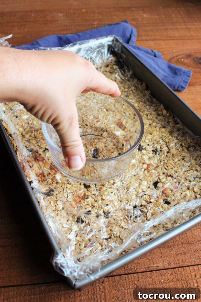 The process of pressing down firmly on homemade granola bars with a bowl over plastic wrap to ensure they are compact and hold their shape.