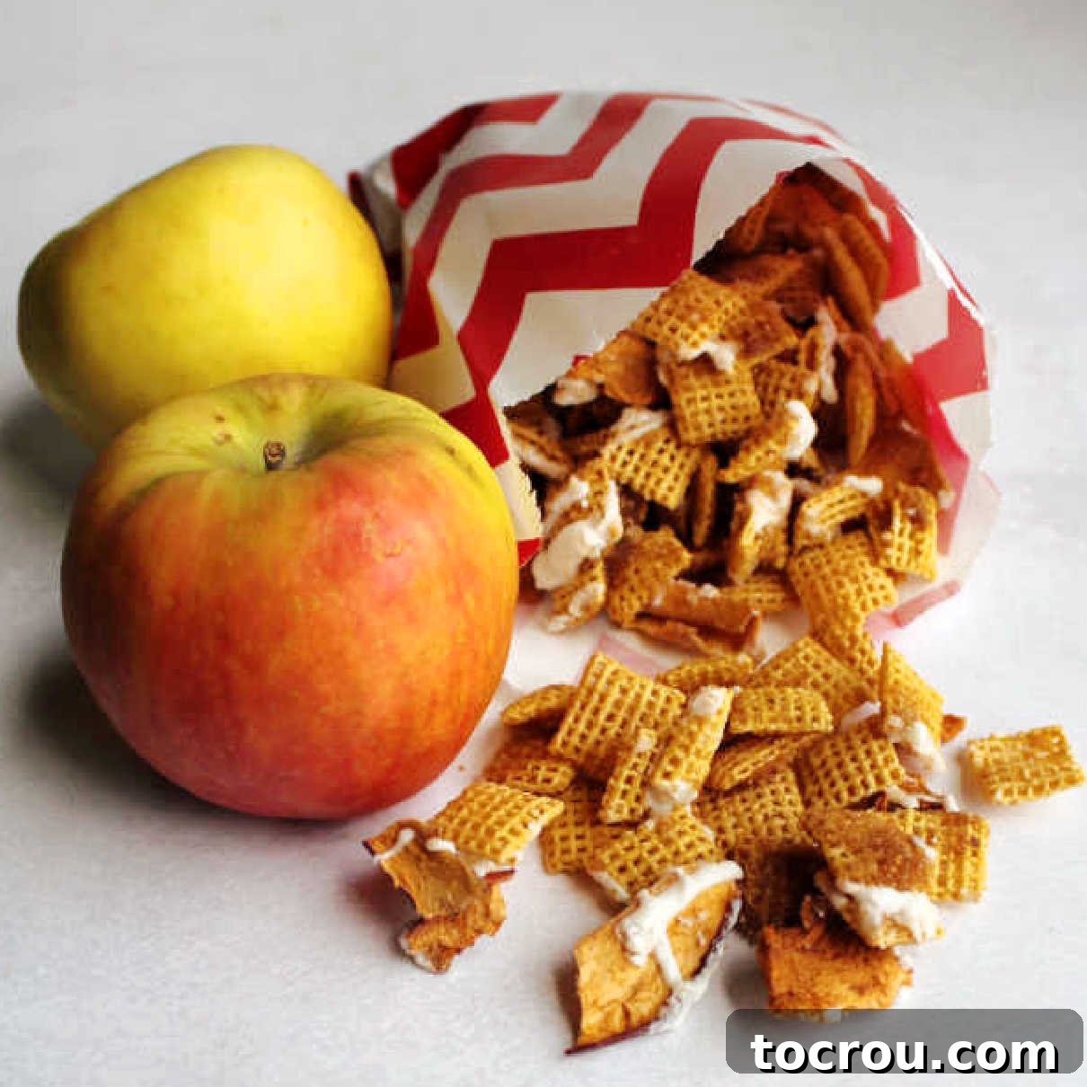 Two apples next to bag of apple cinnamon chex mix with chunks of dried apples and white chocolate drizzle.