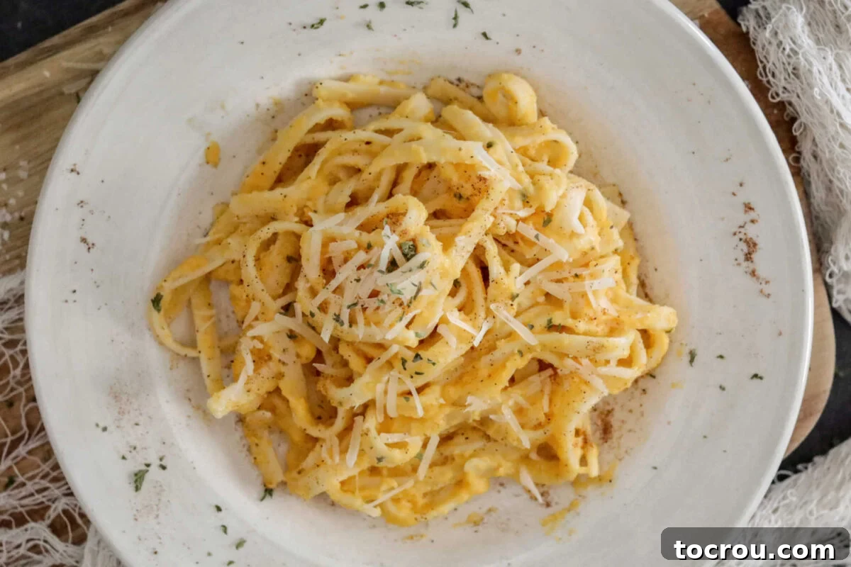 Plate of fettuccine pasta coated in pale orange butternut squash alfredo sauce topped with parmesan cheese, nutmeg, and fresh herbs.