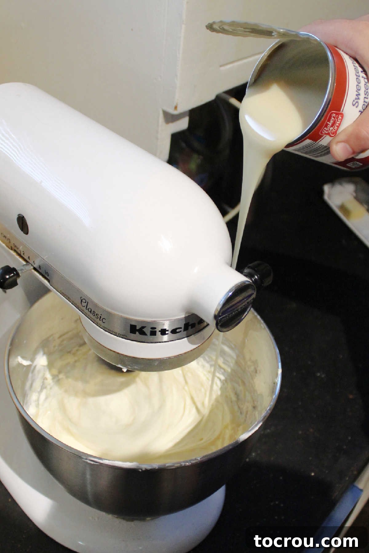 Sweetened condensed milk being slowly poured into a bowl of perfectly beaten, smooth cream cheese, preparing for the next step of cheesecake batter.
