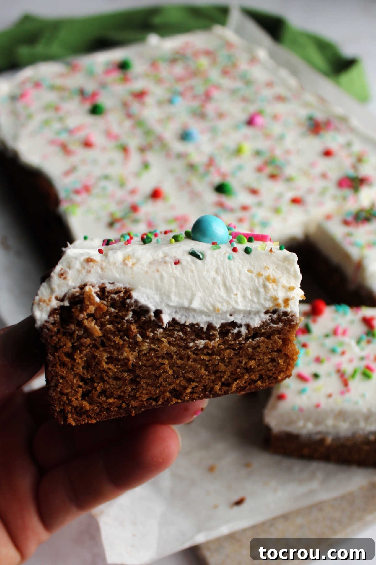 A hand gently holding a thick gingerbread cookie bar, showcasing its soft, chewy texture and the generous layer of fluffy buttercream on top.