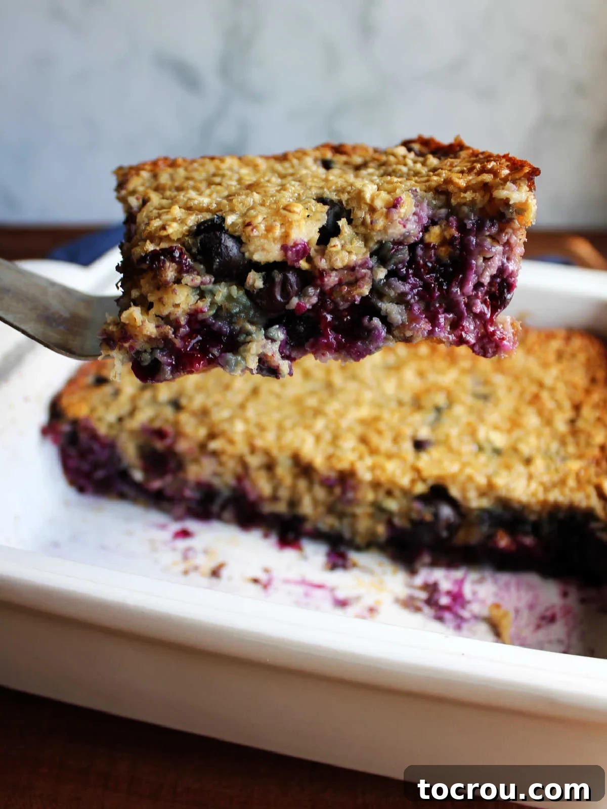 Bright Lemon Blueberry Baked Oatmeal 4 A spatula lifting a perfectly formed piece of lemon blueberry baked oats from a pan, showcasing the abundant fruit and its firm yet tender texture.