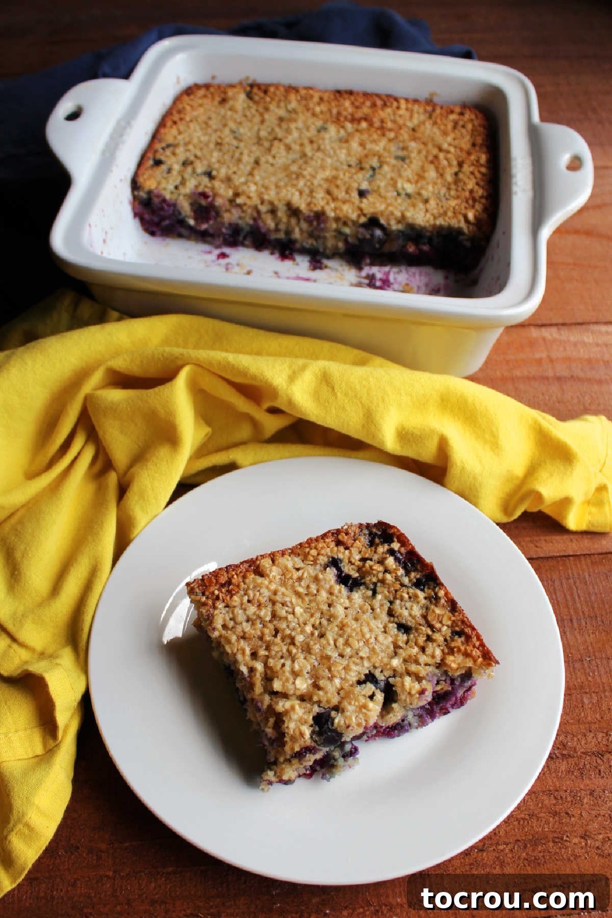 Bright Lemon Blueberry Baked Oatmeal 5 A square slice of lemon blueberry baked oats resting on a small white plate, with a larger square baking dish filled with more baked oatmeal in the soft-focus background.