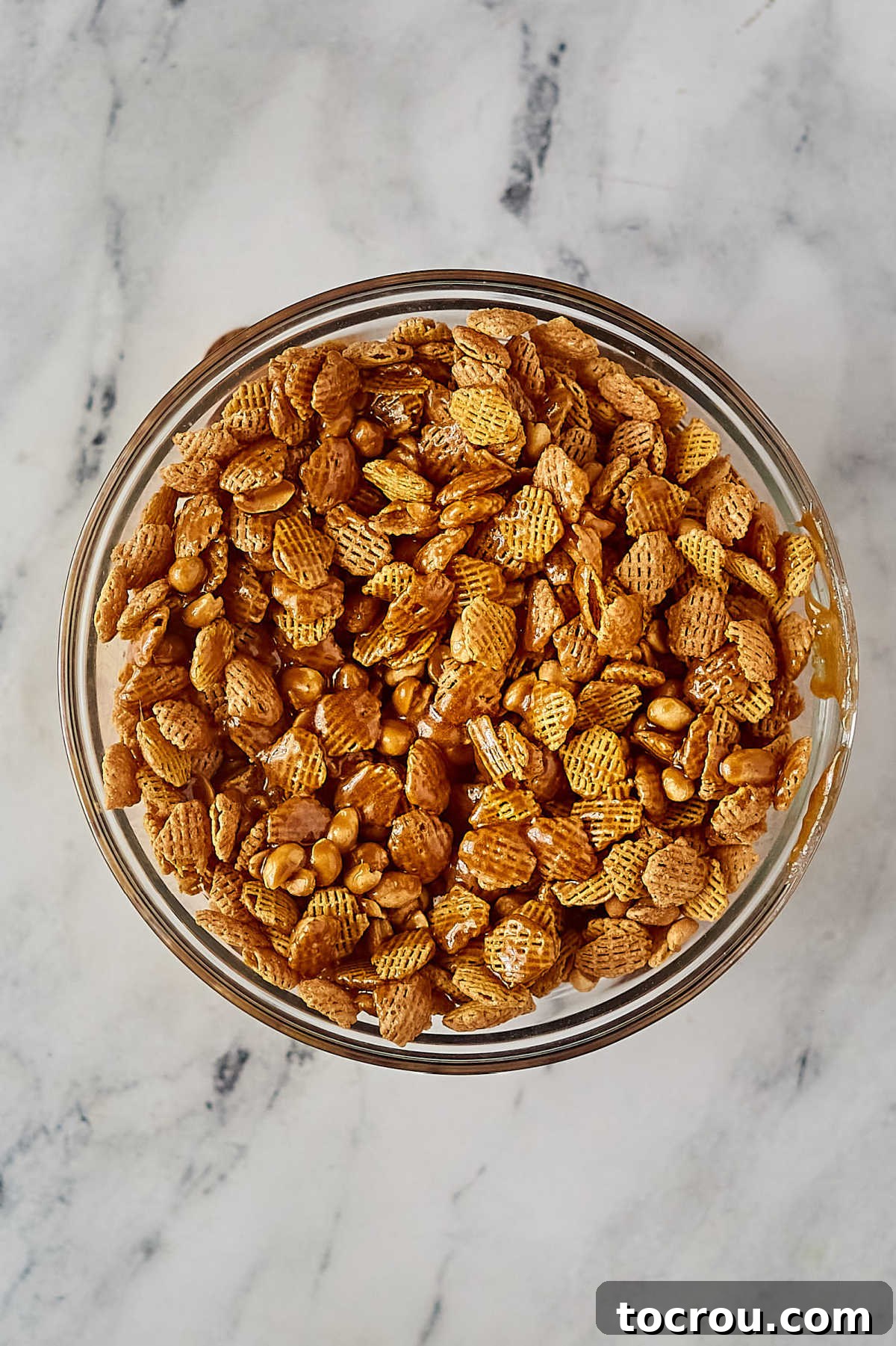 Caramel Crispix in Glass Bowl Glass bowl filled with Crispix cereal, peanuts, coated in hot caramel mixture, ready for microwave steps.