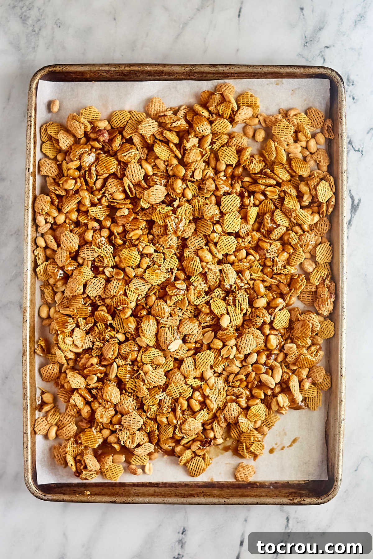 Cooling Caramel Crispix Mix Cooked caramel coated Crispix cereal and peanuts spread out over a wax paper lined baking sheet to cool and set up into individual clusters.